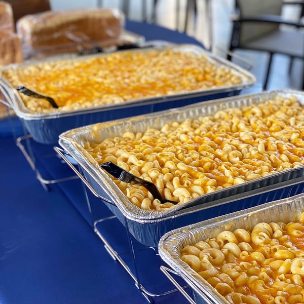 Three foil pans of macaroni and cheese with serving spoons on a blue tablecloth.