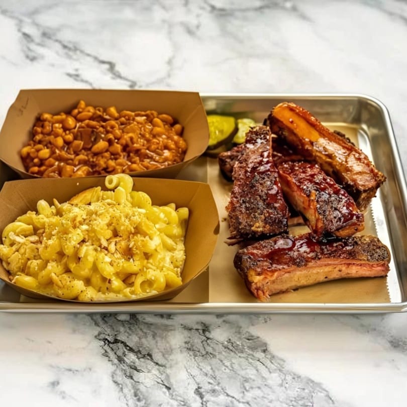 BBQ ribs, baked beans, and macaroni and cheese in paper trays on a metal platter.