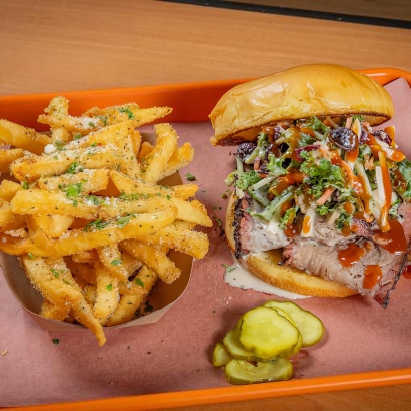 Brisket sandwich with coleslaw and BBQ sauce, a side of seasoned fries, and pickles on an orange tray.
