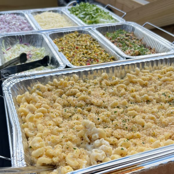 Catering trays filled with macaroni and cheese, coleslaw, corn salad, and other toppings.
