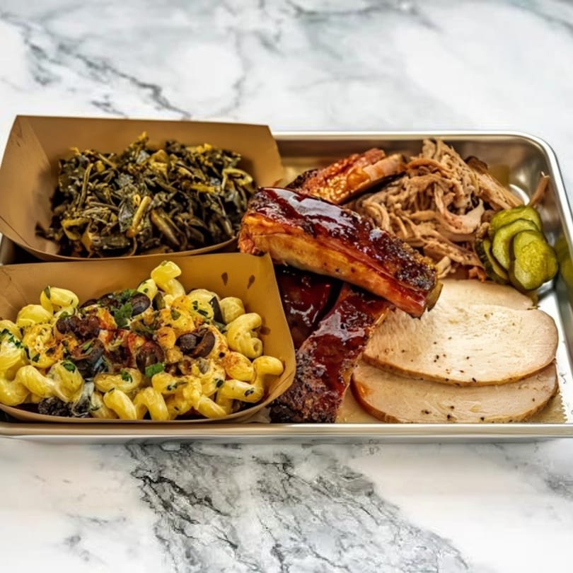 A metal tray with BBQ ribs, pulled pork, sliced turkey, mac and cheese, collard greens, and pickles.