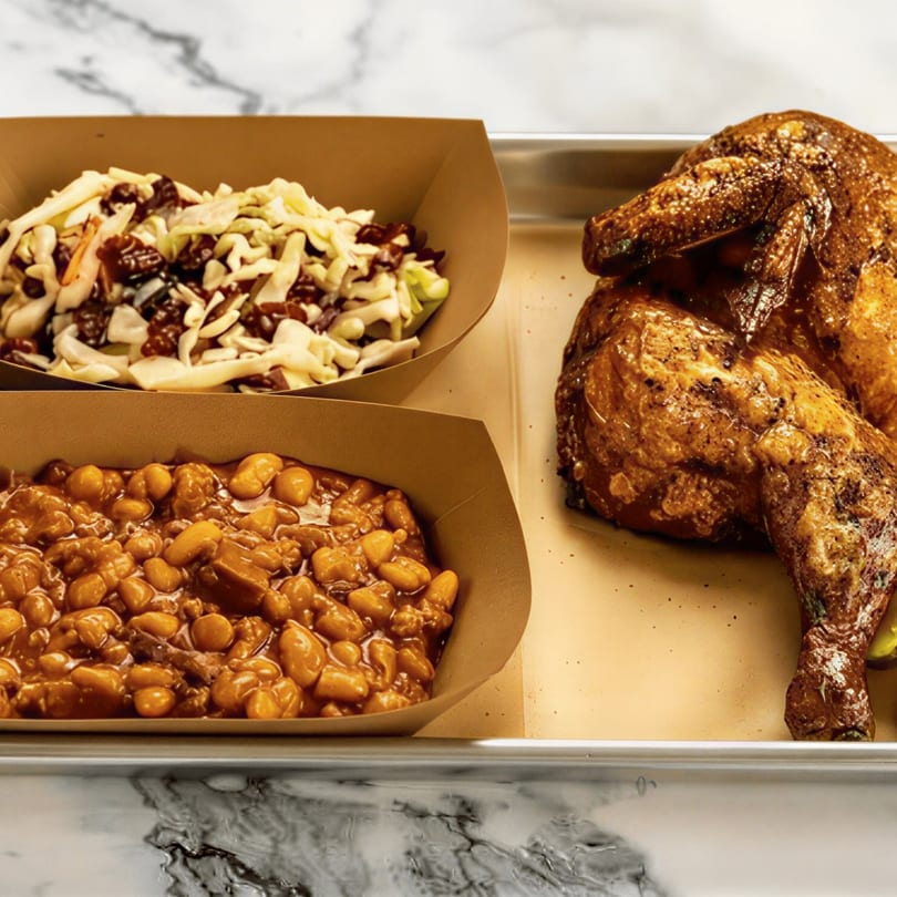 Roasted chicken, baked beans, and coleslaw with cranberries in paper containers on a metal tray.